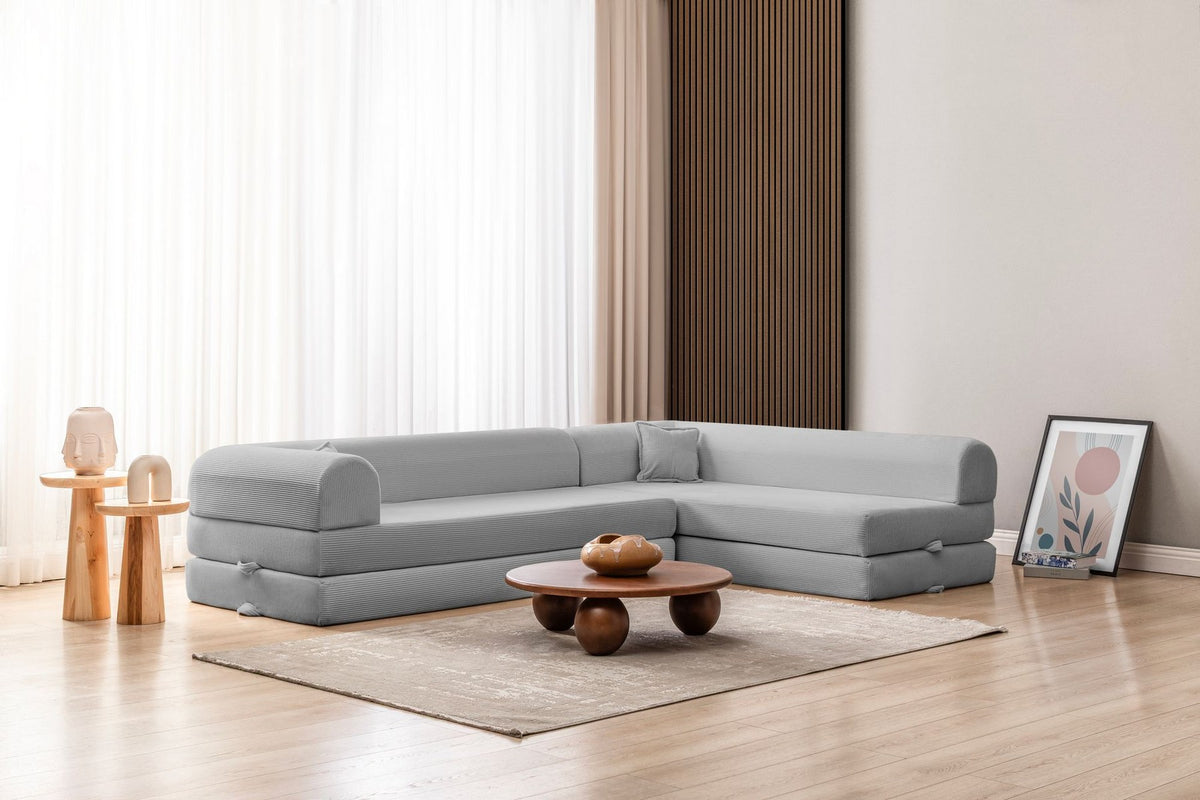Hellgraues Ecksofa-Bett aus geripptem Cord-Plüschstoff mit niedriger Rückenlehne, vier Kissen und ausklappbarer Liegefläche.