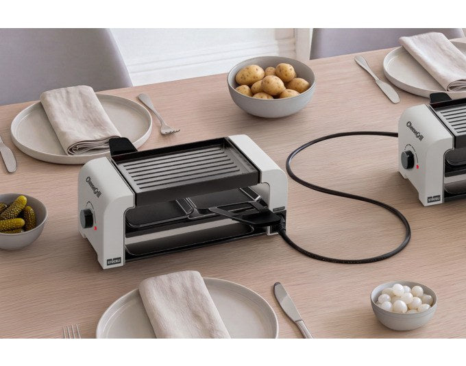 Weisser Tischgrill STÖCKLI CheeseGrill 2 Connect mit wendbarer Aluguss-Grillplatte, zwei Pfännchen und Kunststoffspachtel auf hellem Untergrund.