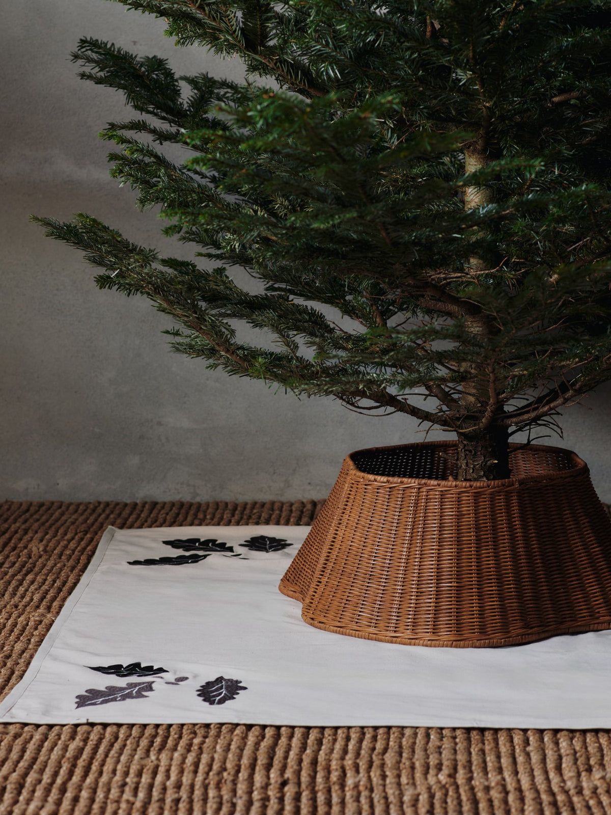 Runde Weihnachtsbaumdecke aus natürlicher Bio-Baumwolle mit aufgestickten Eichenblättern, Beeren und Sternen in dunklem Schokoladenton am Rand.