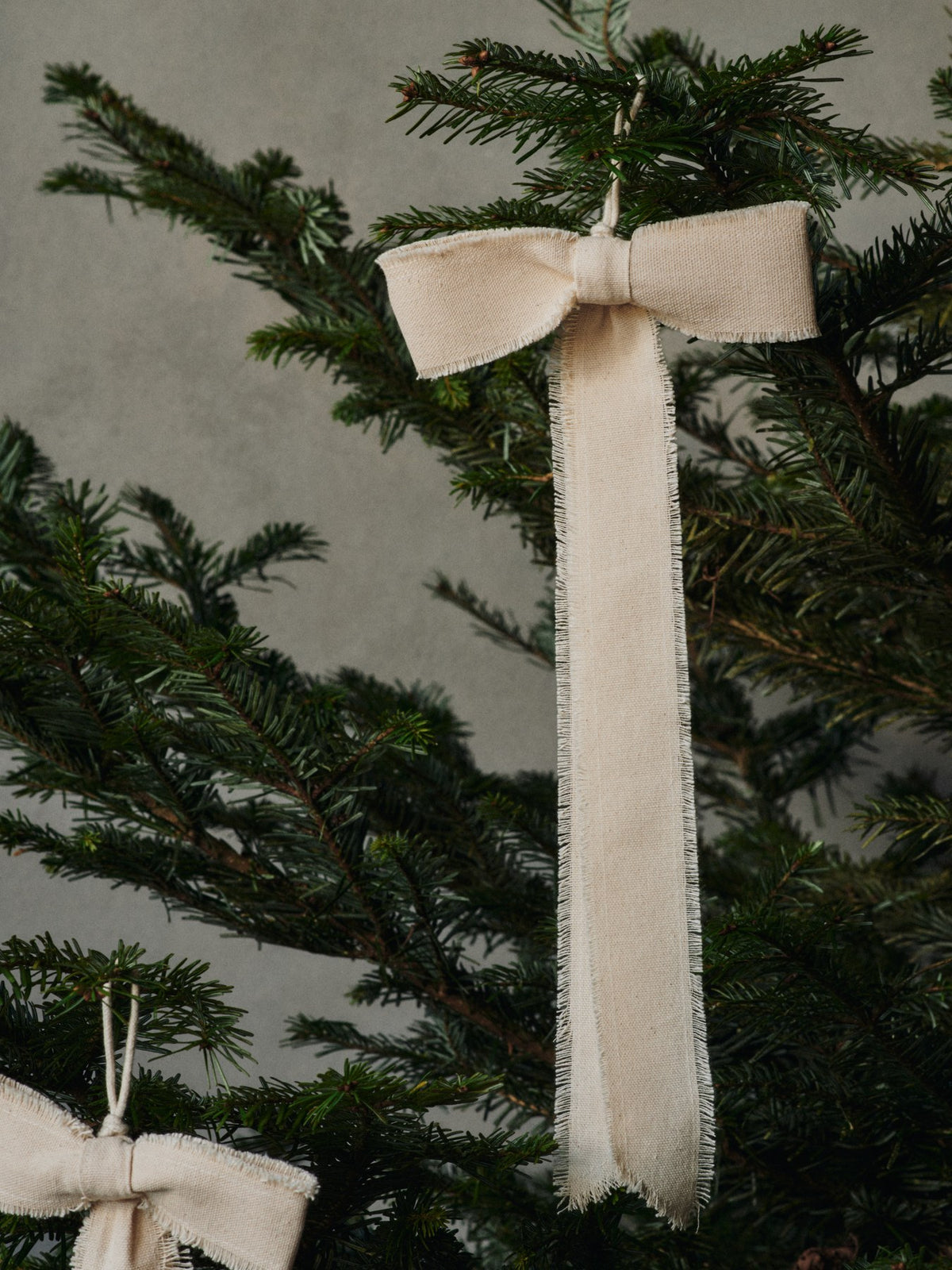 Zwei Dekoschleifen aus Bio-Baumwolle in natürlicher Farbe mit fransigen Kanten und Aufhängeschlaufe, aufgehängt als Weihnachtsschmuck.