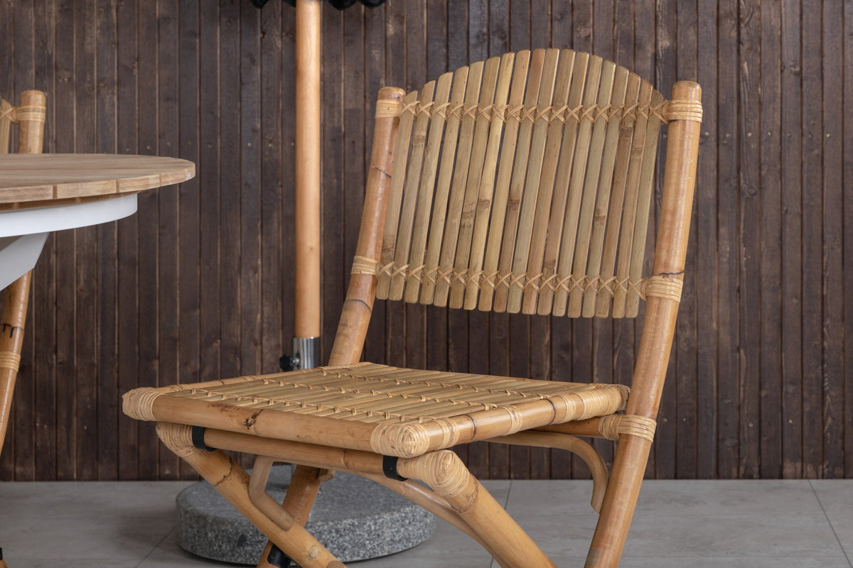 Runder Esstisch ⌀140 aus Teakholz mit Rattanbeinen neben Caféstuhl mit Aluminiumgestell und Bezug aus Baumwolle und Polyester in Naturfarbe.