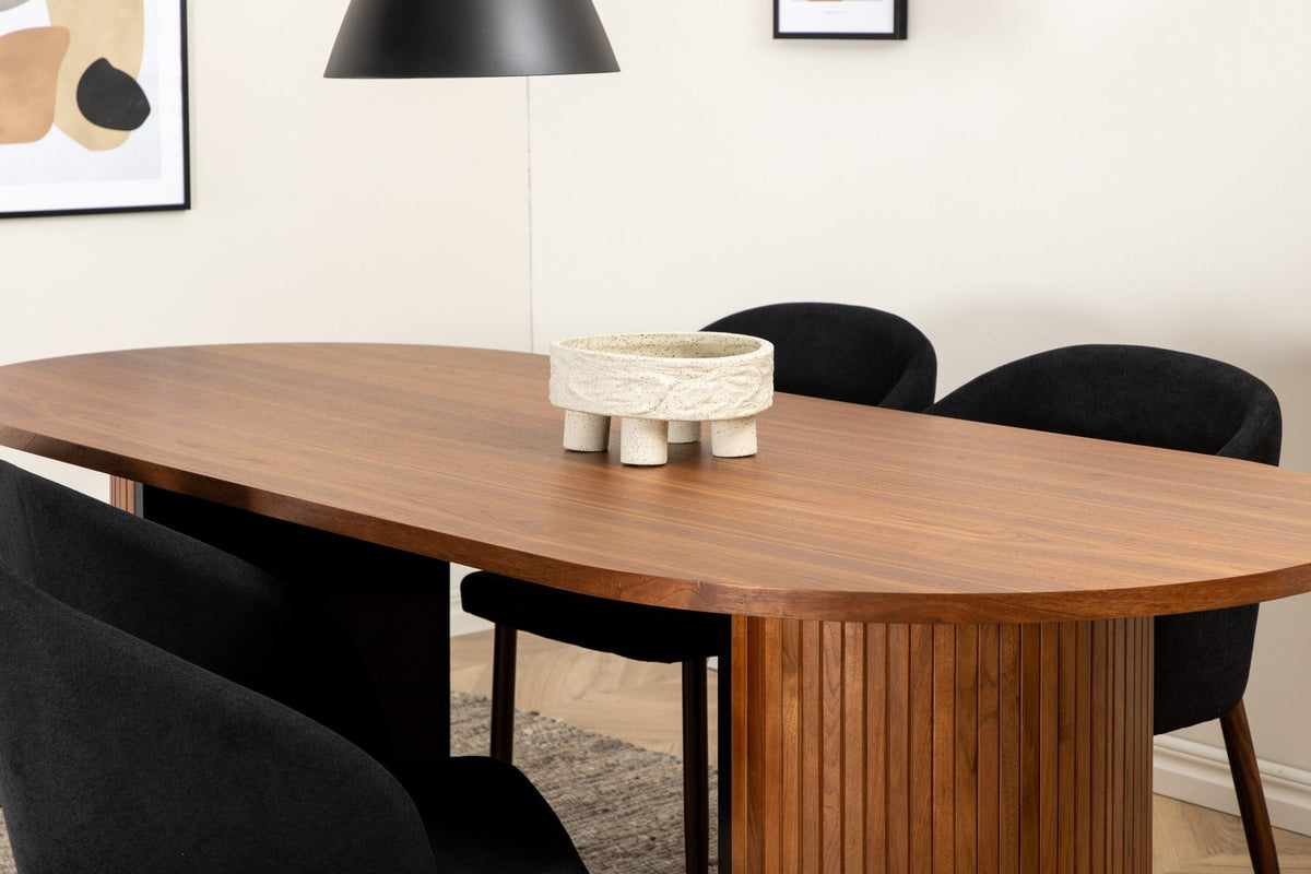 Ovaler Esstisch mit Nussbaum-MDF-Furnierplatte und Stahlgestell, daneben zwei schwarze Polsterstühle mit Metallbeinen im Esszimmer.