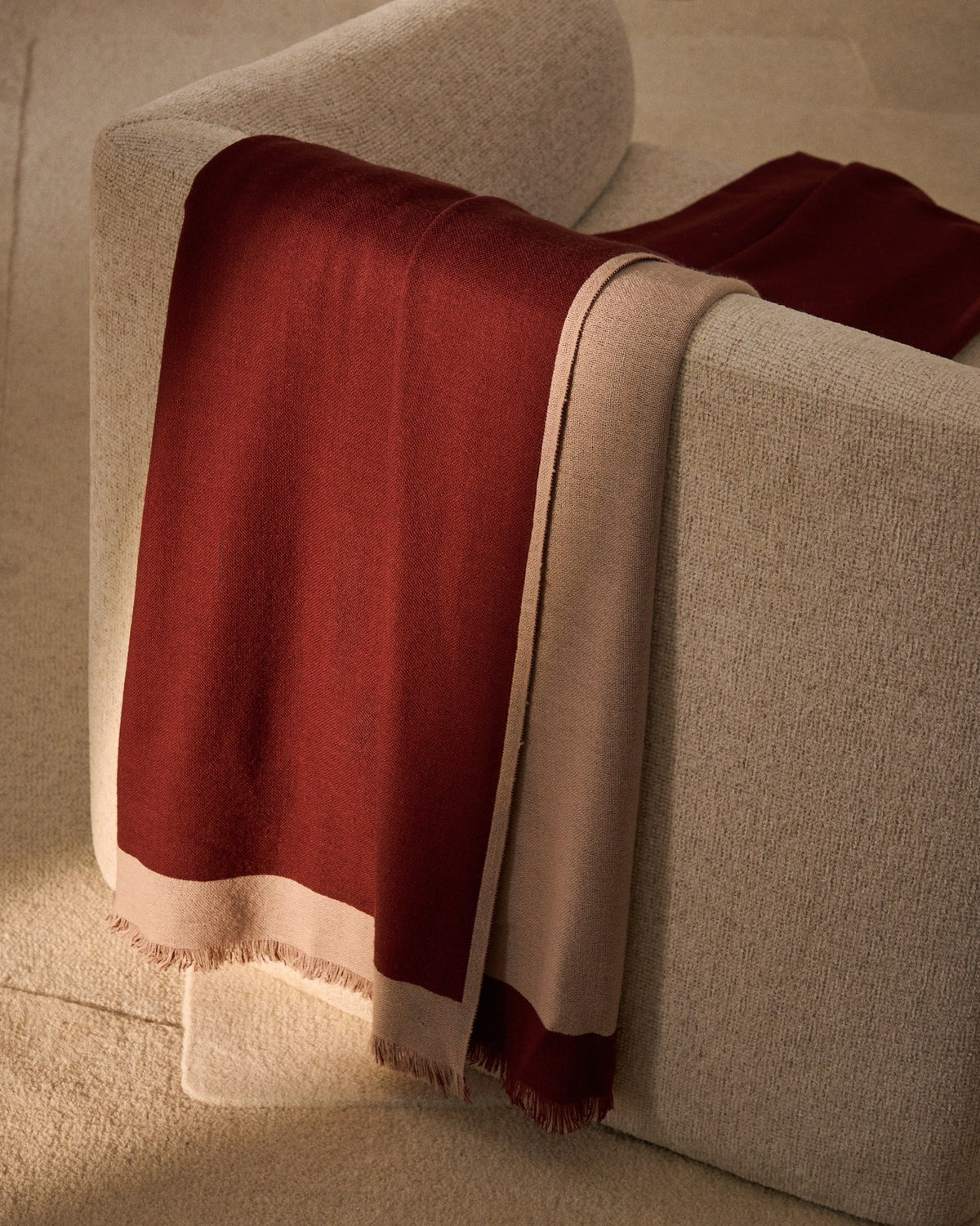 Wendbare Wolldecke aus 100% Wolle in Granatrot und Beige mit Fransen an den Rändern, gefaltet auf einem Sofa liegend.