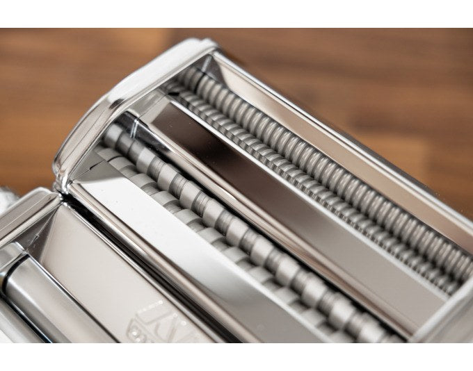 Silberfarbene Nudelmaschine aus Edelstahl mit eloxierten Aluminiumwalzen und Motorantrieb, integrierter Regler für Teigstärke sichtbar.