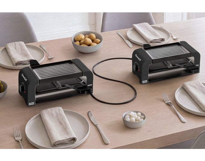 Zwei schwarze STÖCKLI CheeseGrill 2 Connect Tischgrillgeräte mit wendbarer Aluguss-Grillplatte, vier Pfännchen und Kunststoffspachteln.