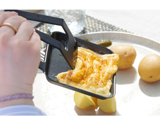 Schwarzer STÖCKLI Tischgrill CheeseGrill 4 Connect mit wendbarer Grillplatte, vier Pfännchen und Kunststoffspachtel auf hellem Untergrund.