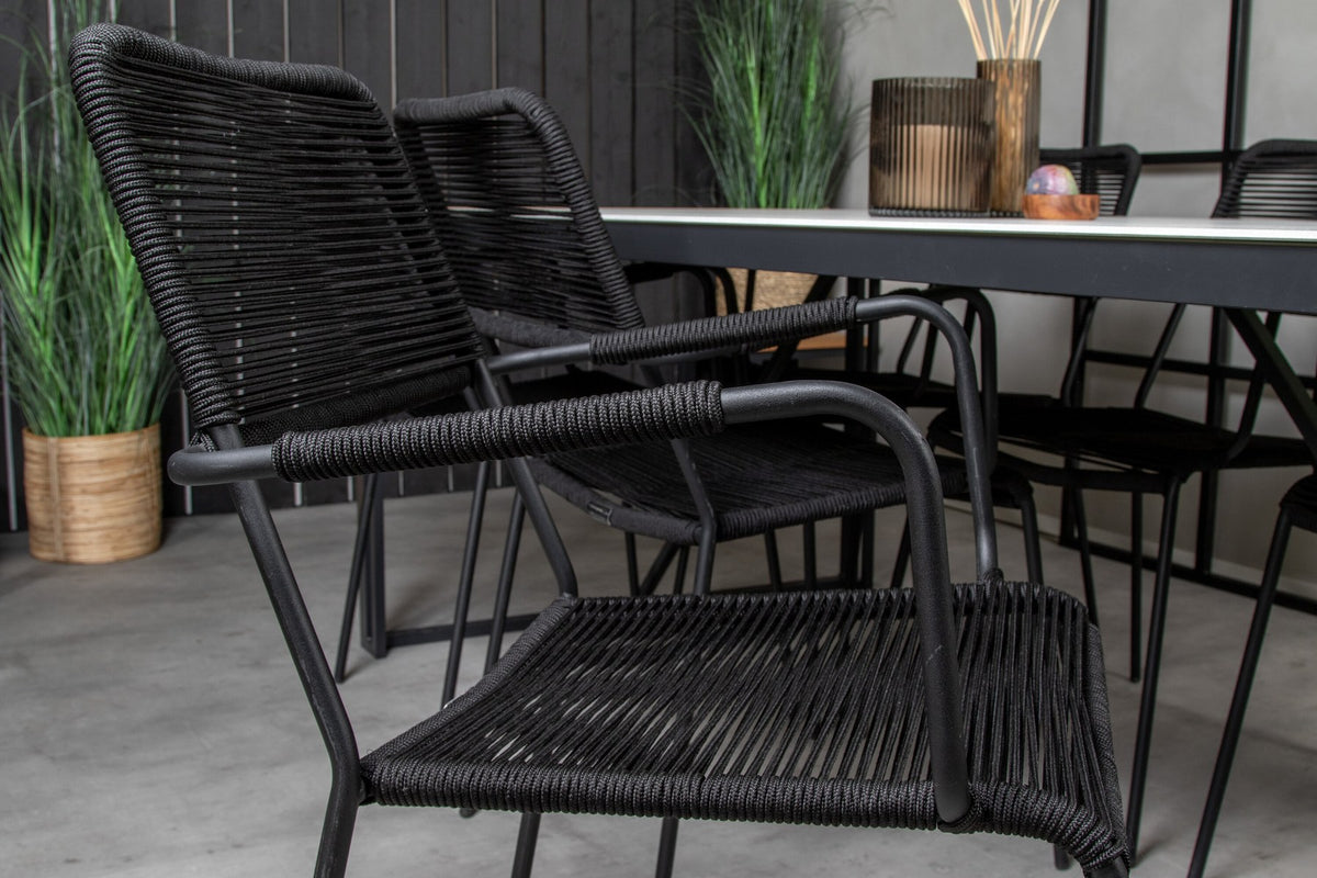 Zwei schwarze Esszimmerstühle mit geflochtener Seilsitzfläche und Aluminiumgestell neben rechteckigem Esstisch mit Glas-Teak-Platte und Stahlbeinen.