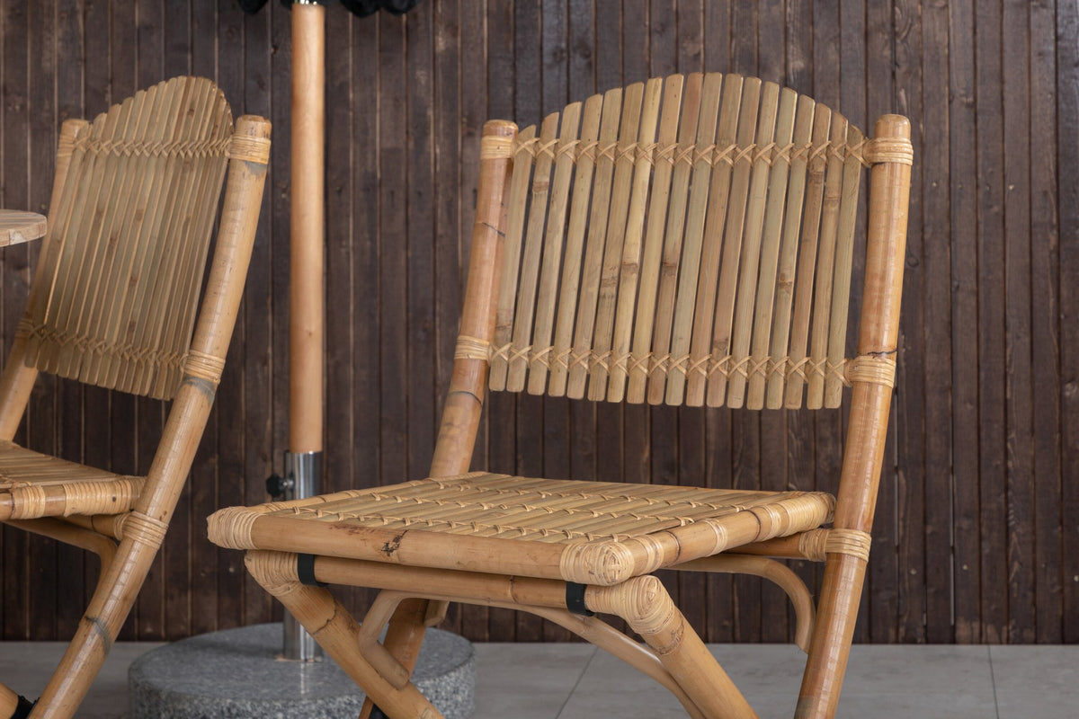 Runder Esstisch ⌀140 aus Teakholz mit Rattanbeinen neben Caféstuhl mit Aluminiumgestell und Bezug aus Baumwolle und Polyester in Naturfarbe.