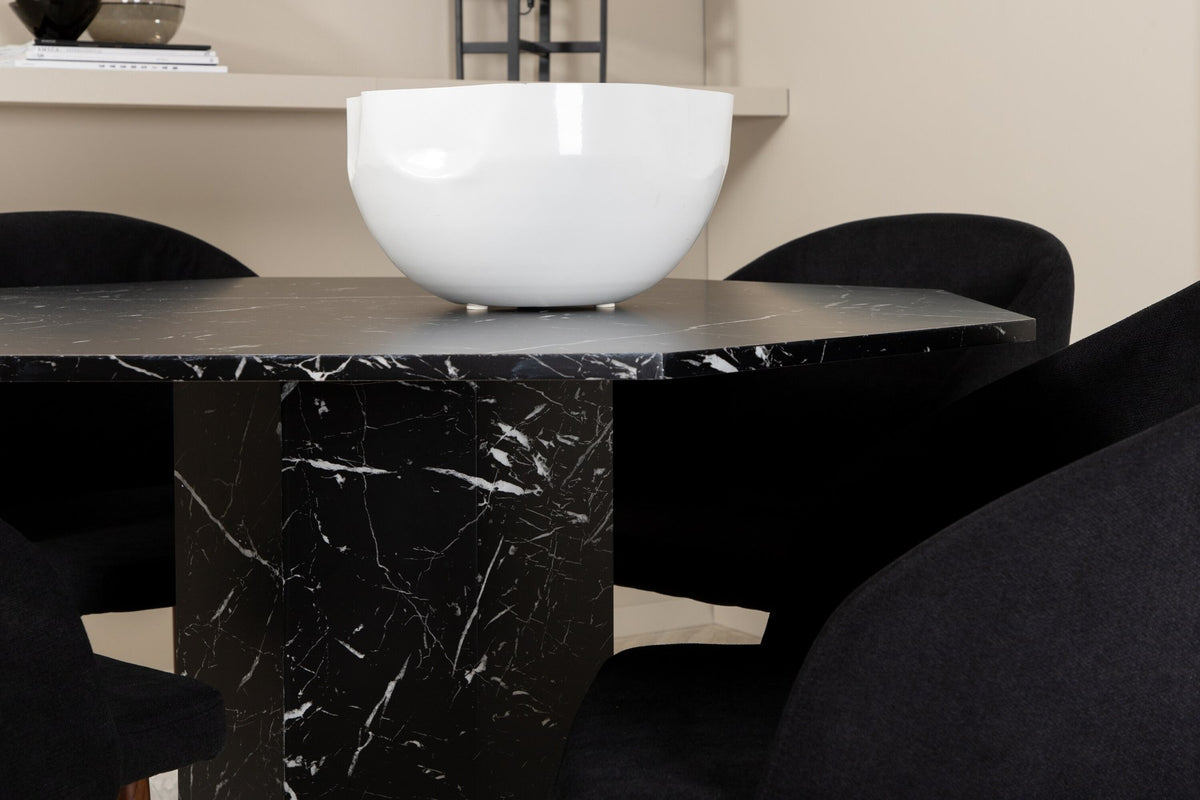 Zwei schwarze Esszimmerstühle mit Nussbaum-Beinen und Polyester-Sitzfläche neben rundem Esstisch Ø110cm mit schwarzer MDF-Platte und Stahlbeinen.