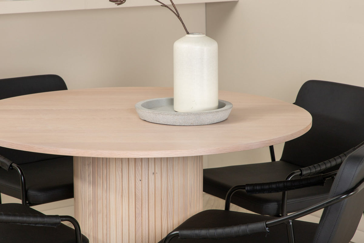 Rundes Esszimmerset aus MDF: Tischplatte Ø110 cm in Whitewash mit gerippten Beinen, dazu zwei schwarze Stühle mit Eisenbeinen.