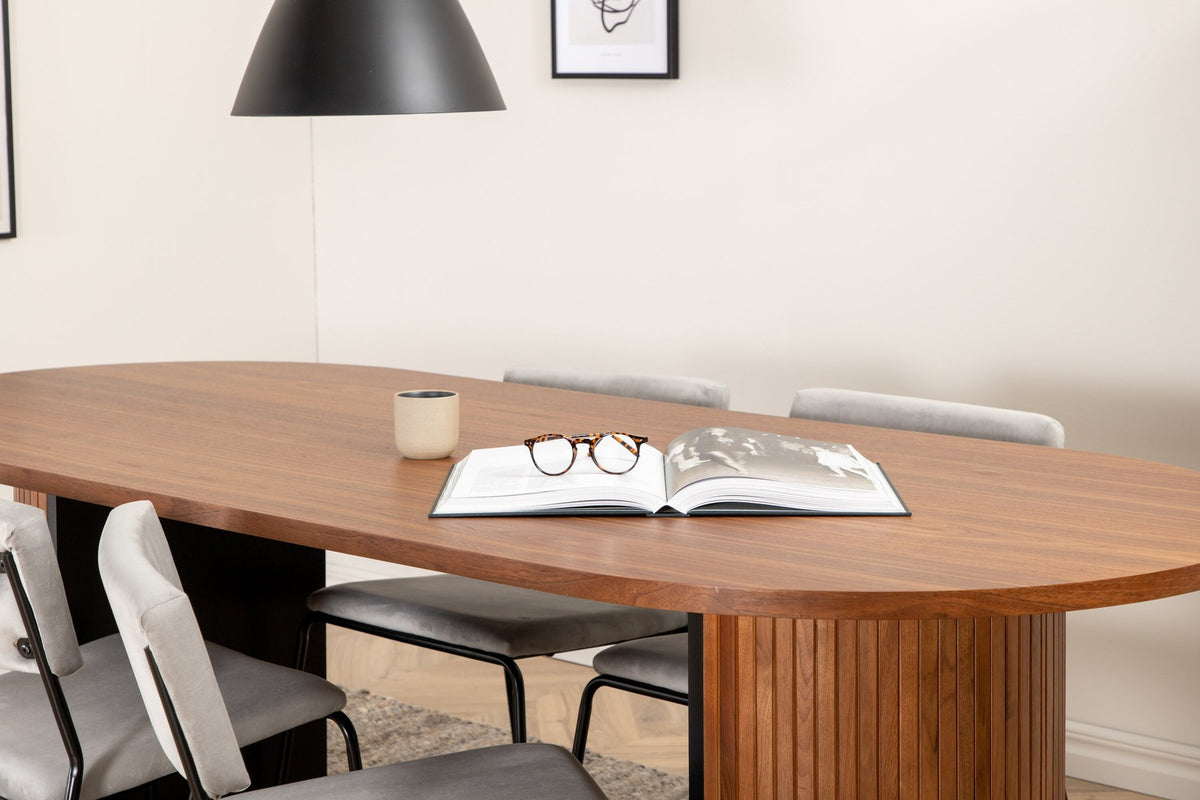 Zwei graue Esszimmerstühle mit schmalen Metallbeinen und samtigem Sitz neben ovalem Esstisch aus Nussbaum-MDF mit Furnier, 200x90cm.