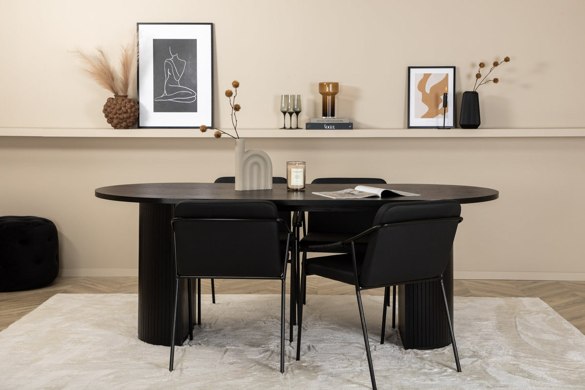 Zwei schwarze Esszimmerstühle mit Beinen aus Eisen und Sitzfläche aus Polyurethan-Membran neben ovalem MDF-Esstisch in Schwarz.