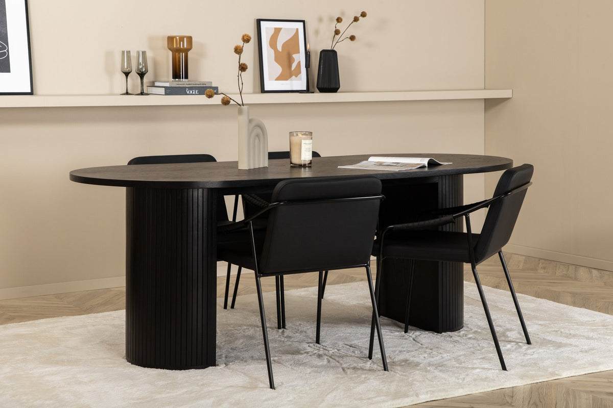 Zwei schwarze Esszimmerstühle mit Beinen aus Eisen und Sitzfläche aus Polyurethan-Membran neben ovalem MDF-Esstisch in Schwarz.