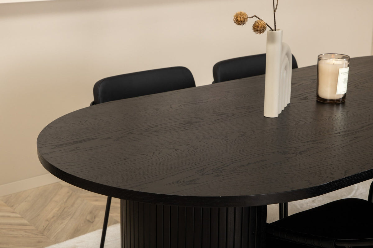 Zwei schwarze Esszimmerstühle mit Beinen aus Eisen und Sitzfläche aus Polyurethan-Membran neben ovalem MDF-Esstisch in Schwarz.