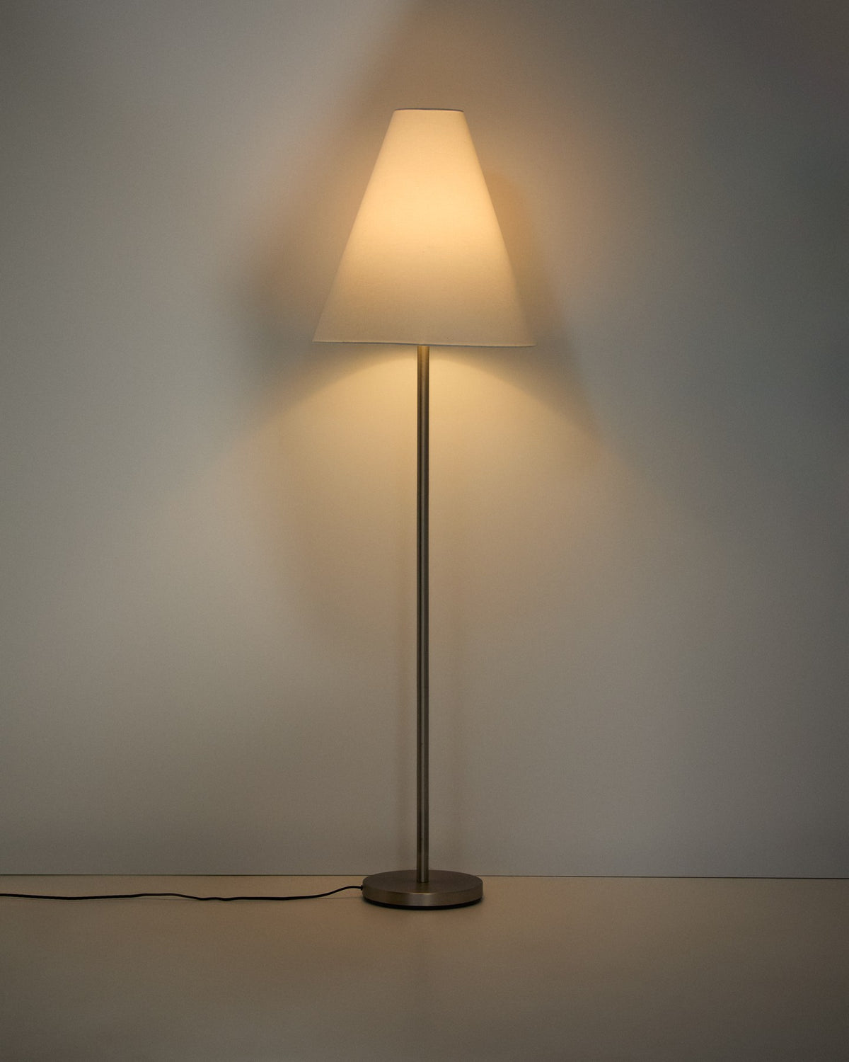 Stehlampe aus Metall mit silbernem, gebürstetem Finish und konischem Lampenschirm aus Baumwollstoff in gebrochenem Weiss, mit Stahlsockel.