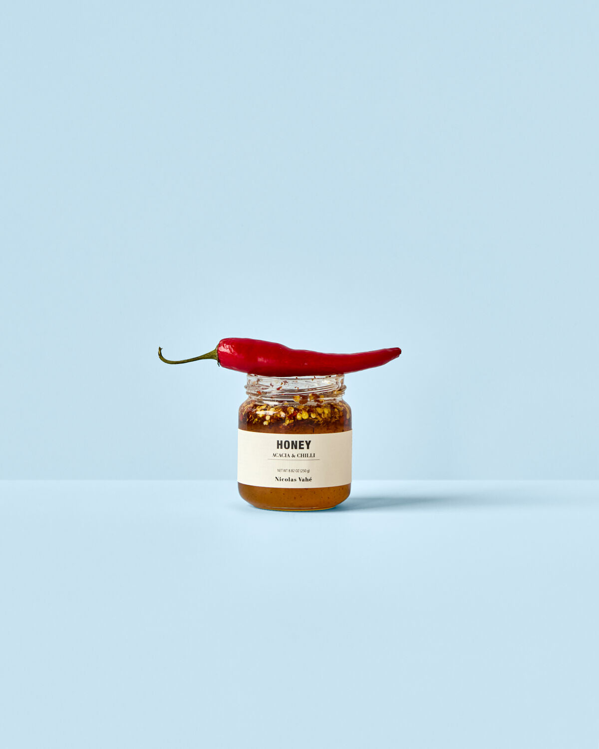 Ein Glas mit Honig, Akazie und Chili von Nicolas Vahé, 250 g, stilvoll präsentiert auf einem rustikalen Holzuntergrund.