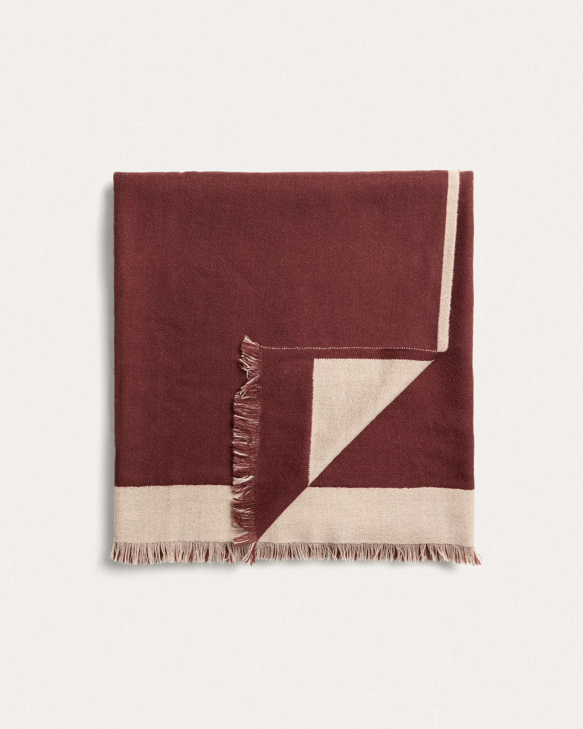 Wendbare Wolldecke aus 100% Wolle in Granatrot und Beige mit Fransen an den Rändern, gefaltet auf einem Sofa liegend.