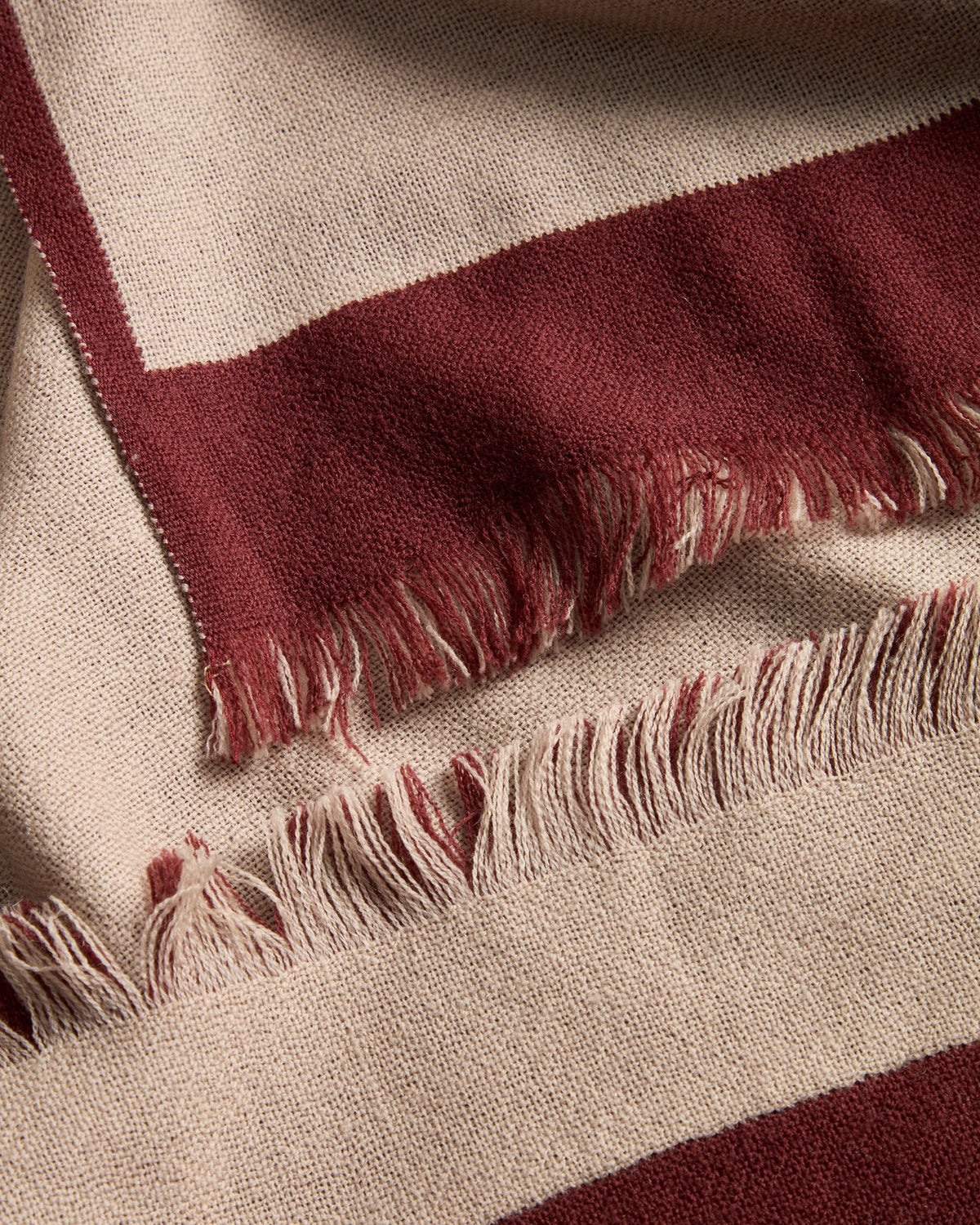 Wendbare Wolldecke aus 100% Wolle in Granatrot und Beige mit Fransen an den Rändern, gefaltet auf einem Sofa liegend.